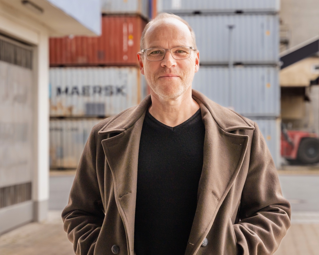 Ein Mann mit Brille blickt in die Kamera. Er steht in einem Containerhafen