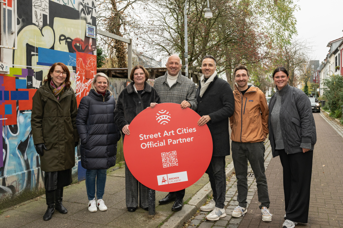 Seven people are standing on a street holding up a Street Art Cities Official Partner sign