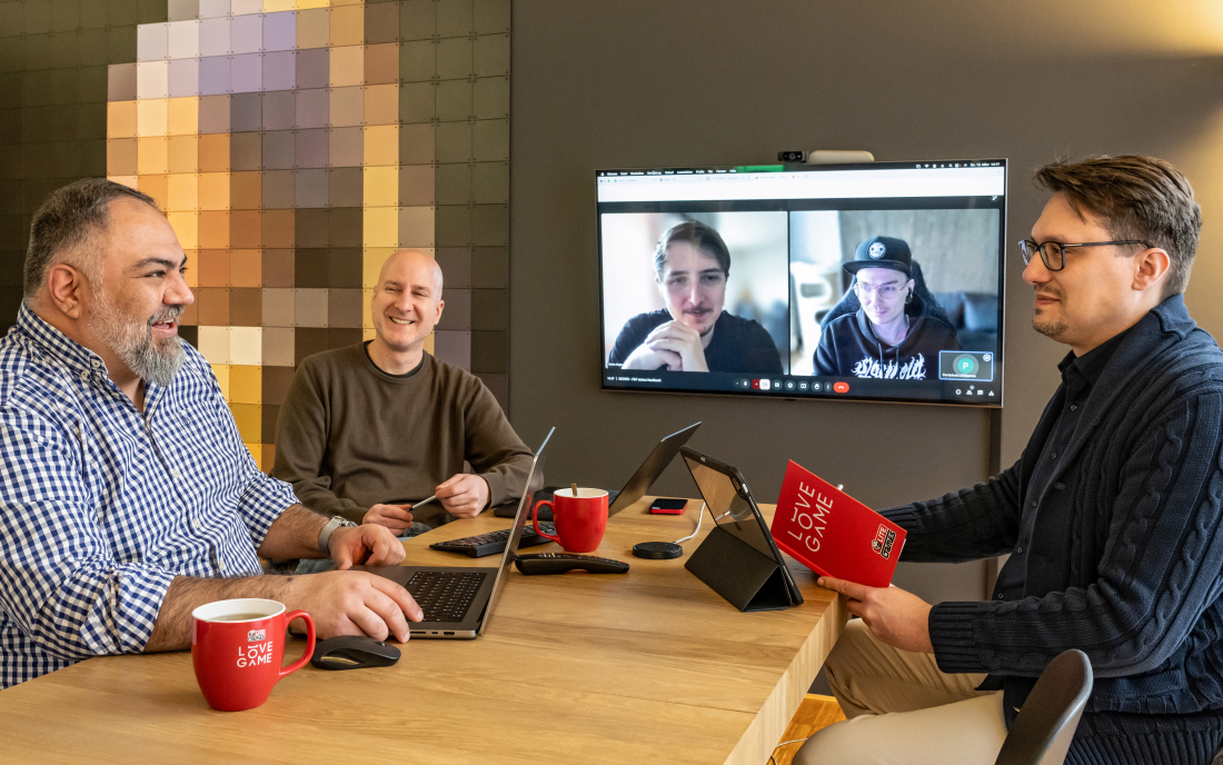 Drei Männer sitzen um einen Tisch. An der Wand hängt ein Bildschirm mit einer Videokonferenz