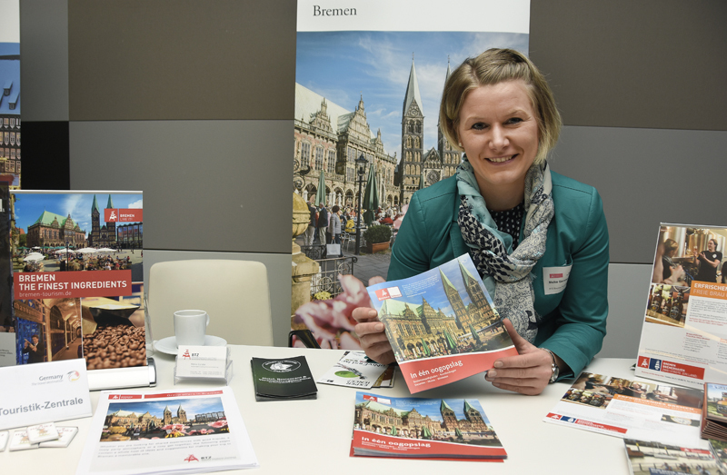 Eine blonde Frau in einem türkisen Blazer sitzt an einem Tisch und hält einen Bremen-Reisekatalog in der Hand