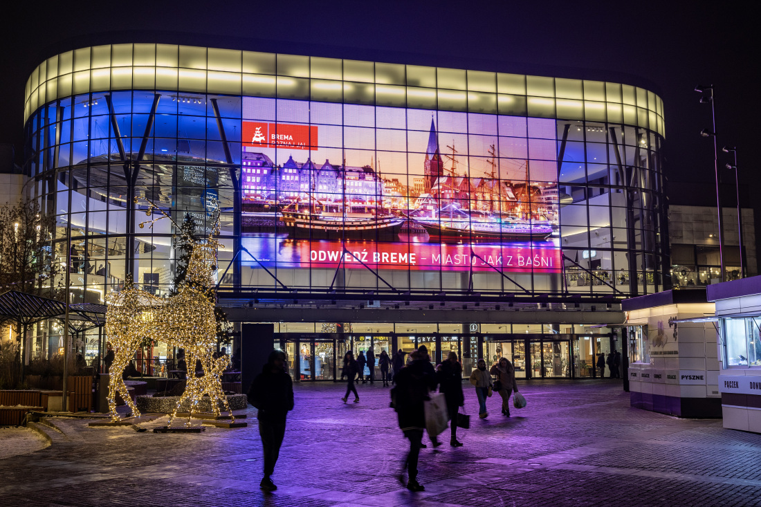 Man sieht ein Einkaufszentrum, in das Menschen gehen und auf dem Gebäude ist ein großer Screen, der Bremen Werbung abbildet.