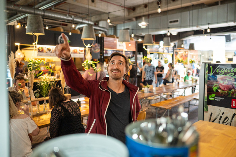 Ein Mann steht in einer belebten Halle mit Biertischen und -Bänken. Um ihn herum stehen und sitzen Menschen. Er zeigt auf etwas, das oben hinter dem Betrachtenden steht.