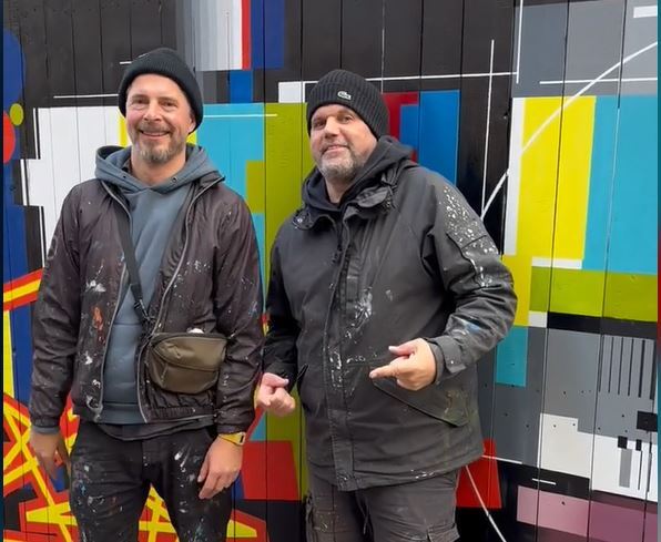 Two men with black caps and clothes stained with paint from a street art artwork