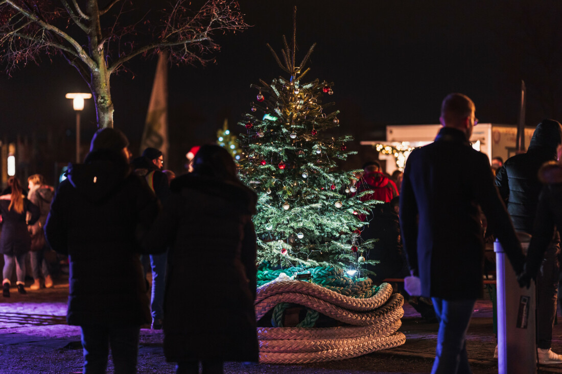 Menschen, die sich im Abendlicht um einen kleinen Platz mit Ständen bewegen. Im Mittelpunkt steht ein geschmückter Weihnachsbaum, der am Fuß mit einem dicken Tampen umlegt ist.