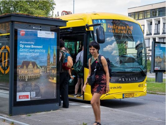 Man sieht eine Bushaltestelle, an der ein Bus steht. Menschen steigen aus und ein. An der Bushaltestelle ist ein Display mit Bremen Werbung. 