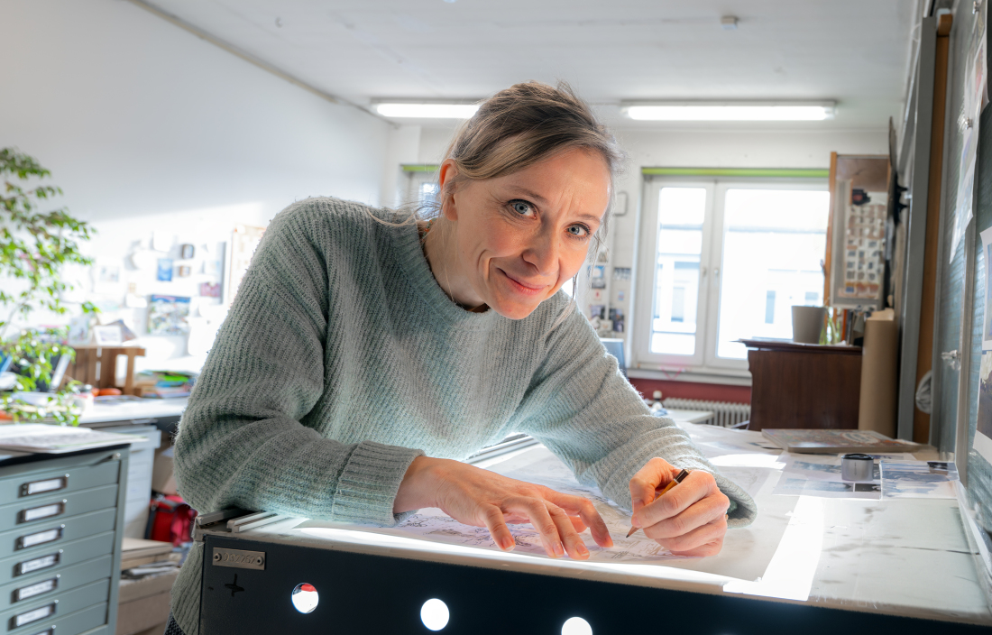 Die Illustratorin Valeska Scholz beugt sich über einen von unten beleuchteten Tisch und zeichnet mit einem Bleistift Figuren auf ein Blatt Papier.