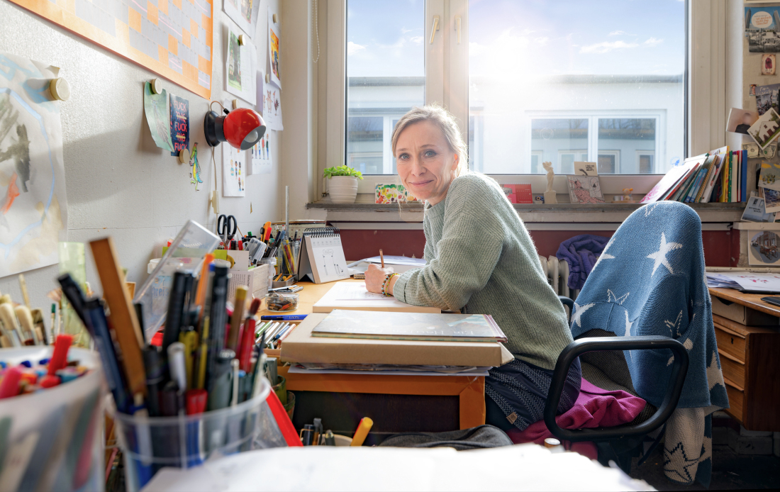 Die Illustratorin Valeska Scholz sitzt an ihrem Schreibtisch und lächelt in die Kamera.